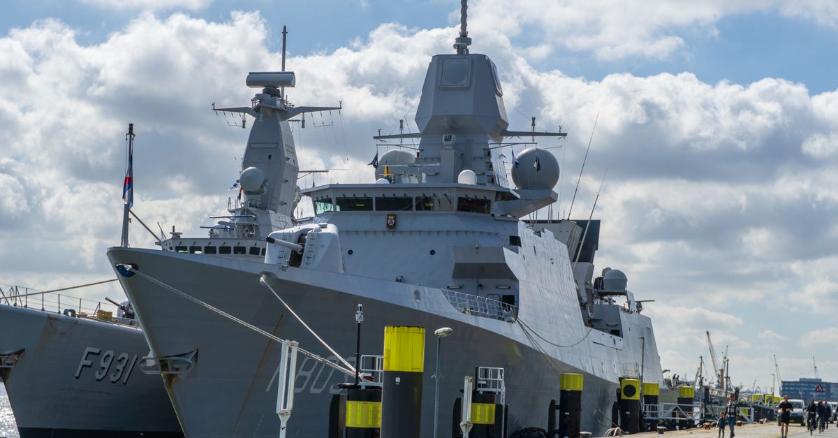 A modern naval warship moored at the port of Rotterdam under a blue sky with clouds.